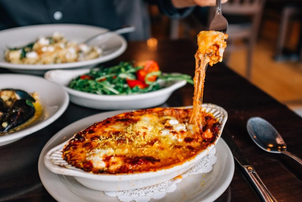 A fork lifts a cheesy bit of food from a white dish on a brown wooden table, surrounded by other dishes of food